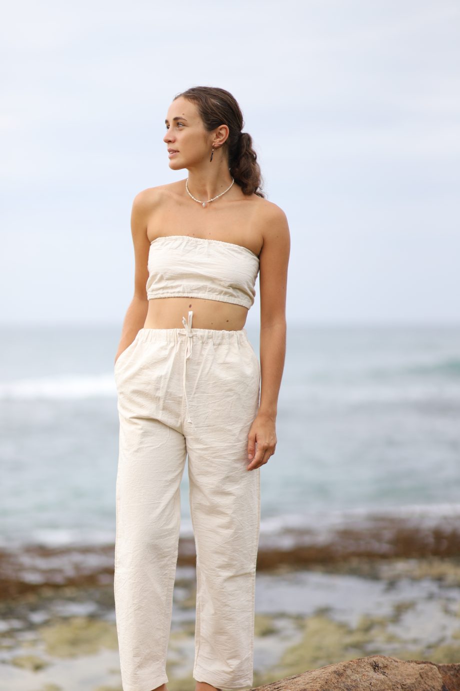 "A front view of a model smiling and posing with one hand raised to her head, wearing the Ethereal Tube Top. The natural off-white hemp fabric highlights the top's strapless design and frilled hem, emphasizing a free-spirited, eco-conscious style