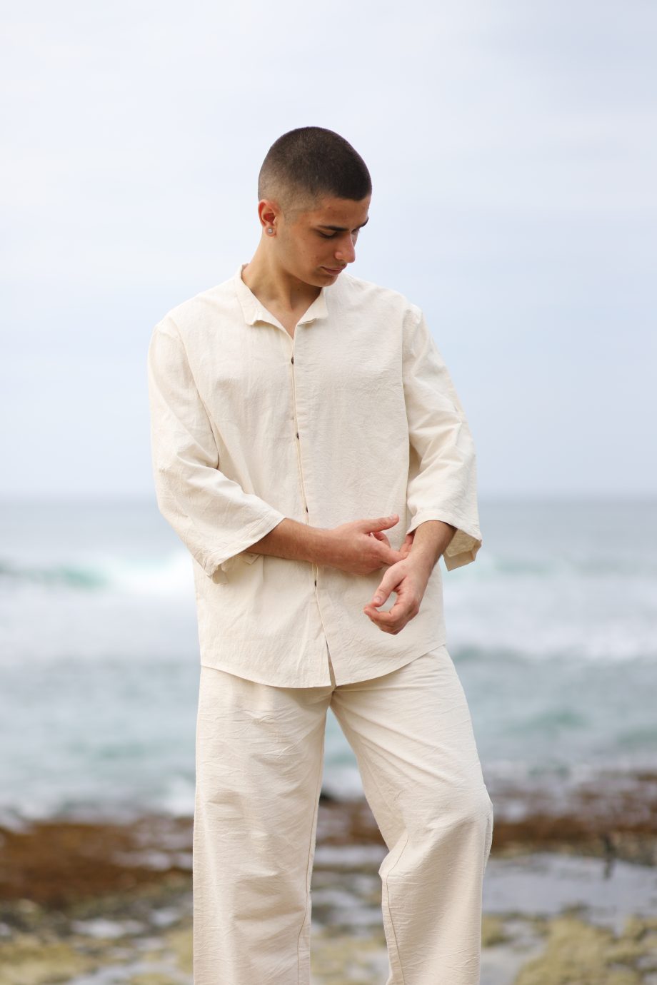 The image features a young man on a beach, exuding casual elegance in the "Tibetan Breeze Shirt" by Higher Himalayan. This off-white, hemp fabric shirt features a button-down front, a soft collar, and a relaxed fit that complements the serene coastal setting.